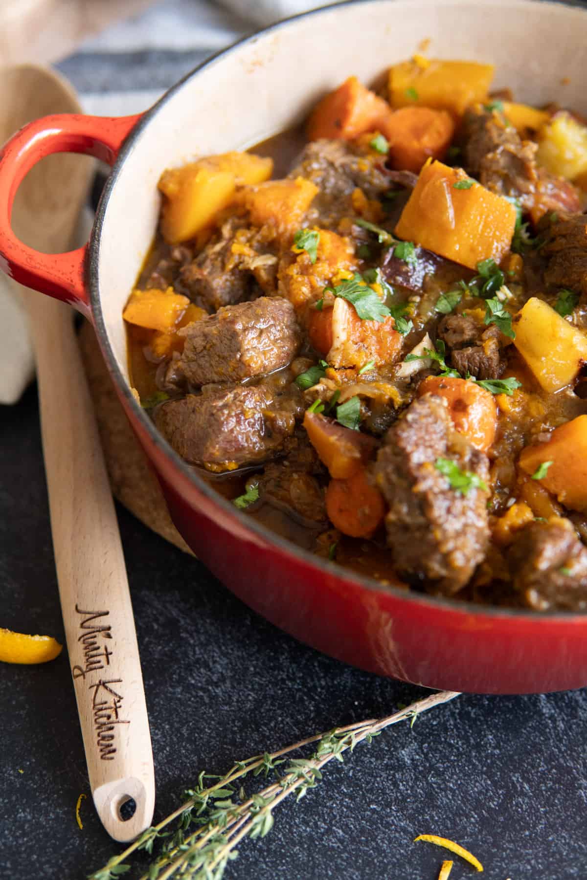 A dutch oven filled with beef stew with squash and carrots and wooden spoon on side. 