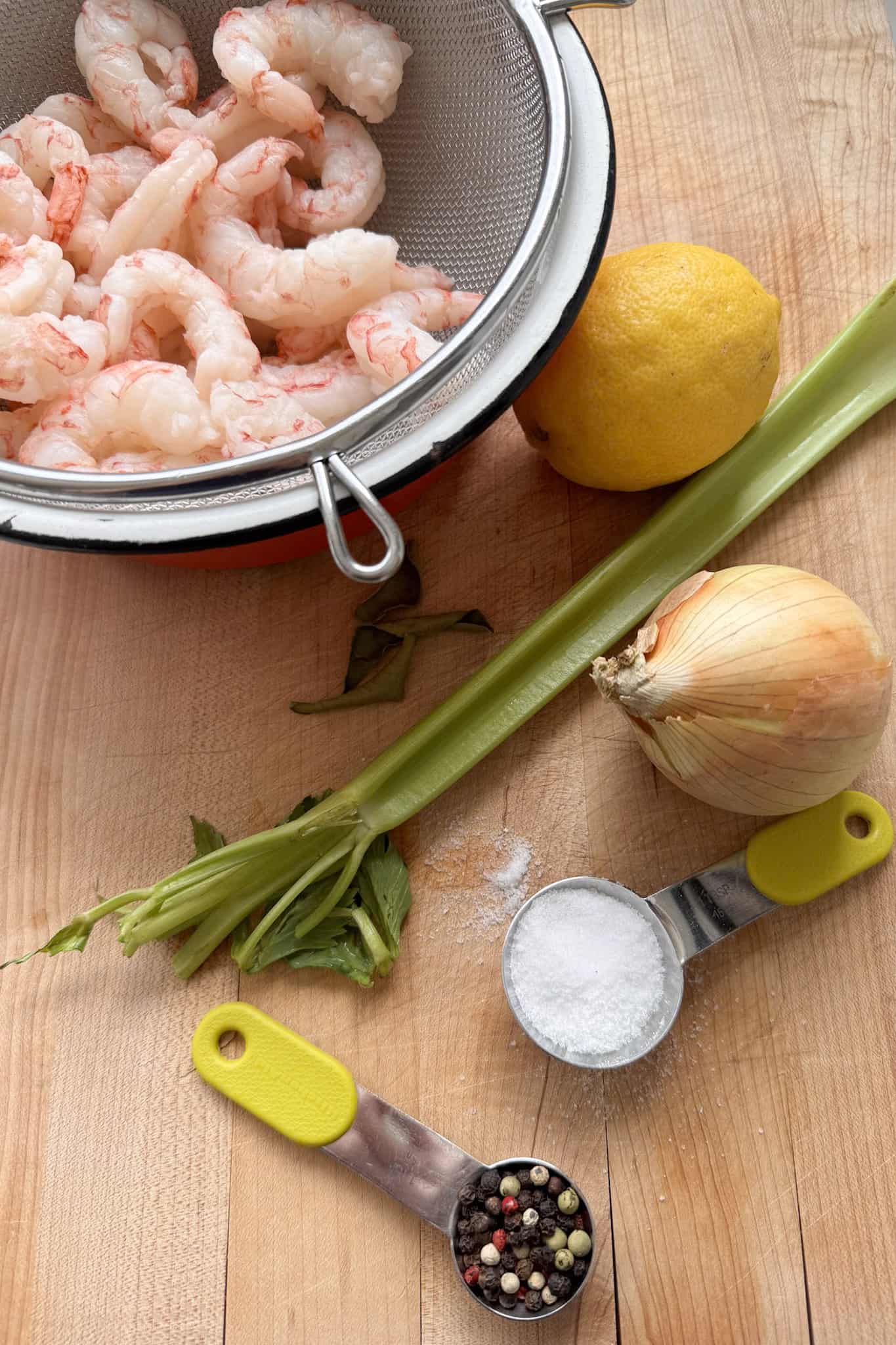 raw shrimp in colander, onion, lemon, celery, salt and peppercorns on cutting board 