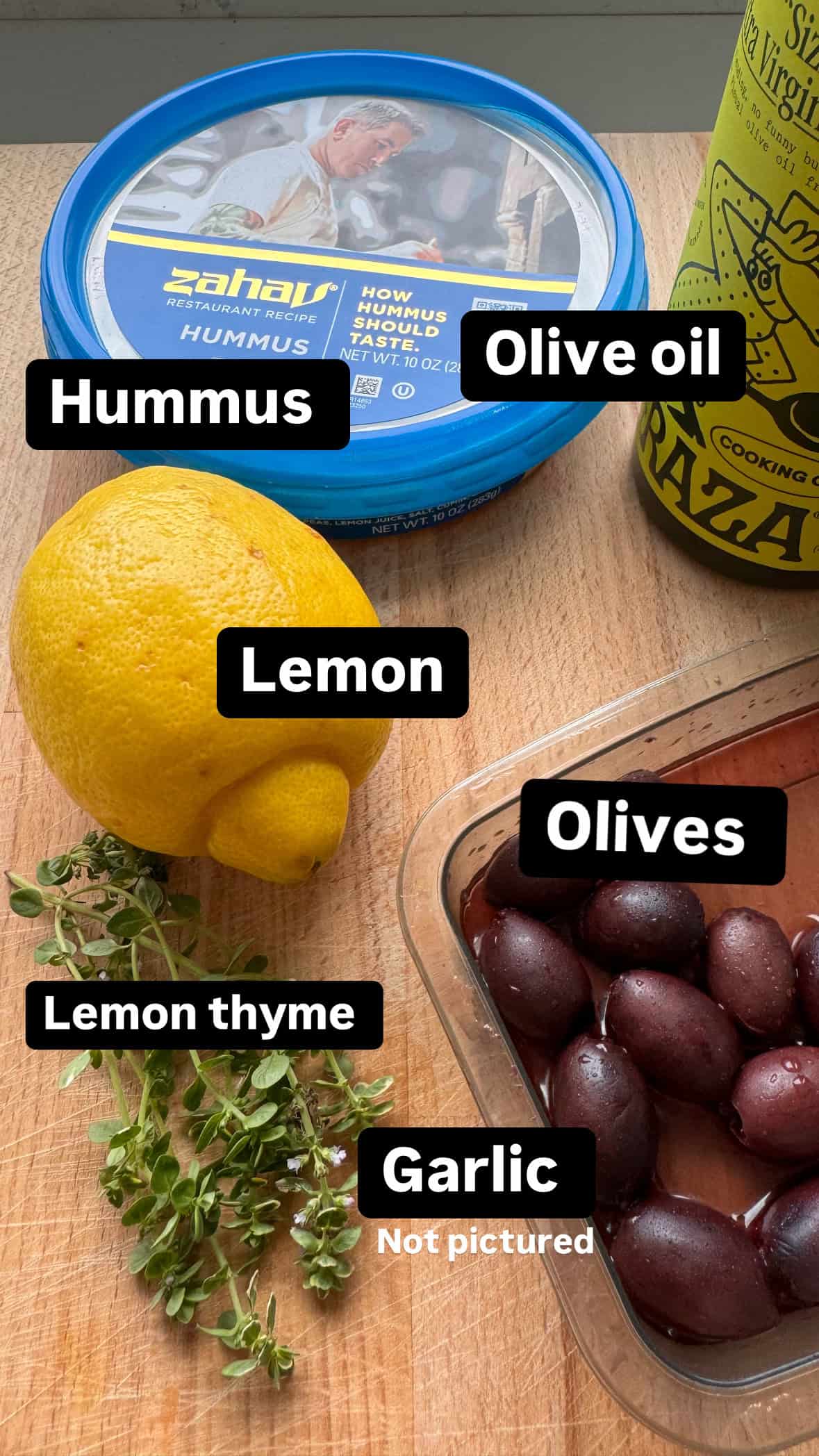 cutting board with hummus container, lemon, olives, thyme and olive oil