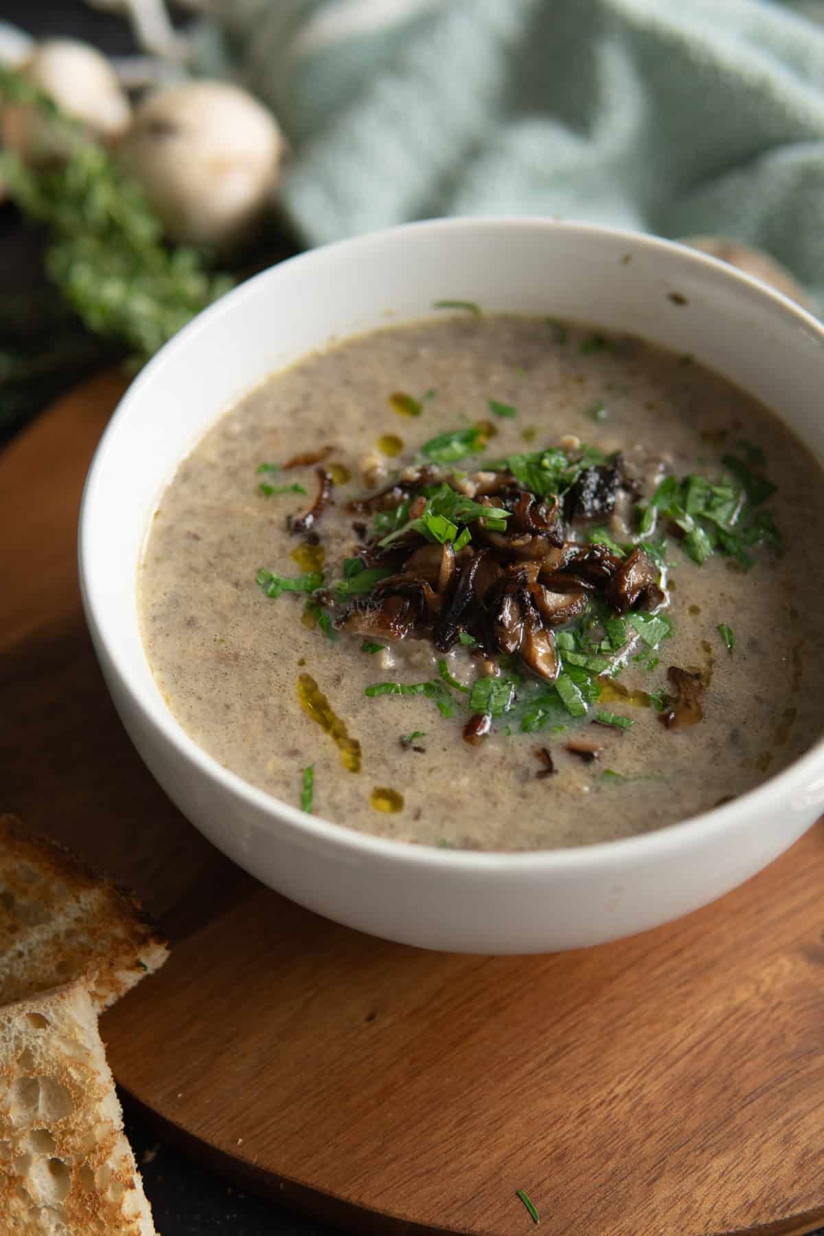 a close up of a white bowl with mushroom soup garnished with seared mushrooms and fresh herbs and a drizzle of olive oil.