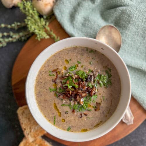 bowl of creamy mushroom soup with spoon next to it, and 2 slices of bread, fresh herbs