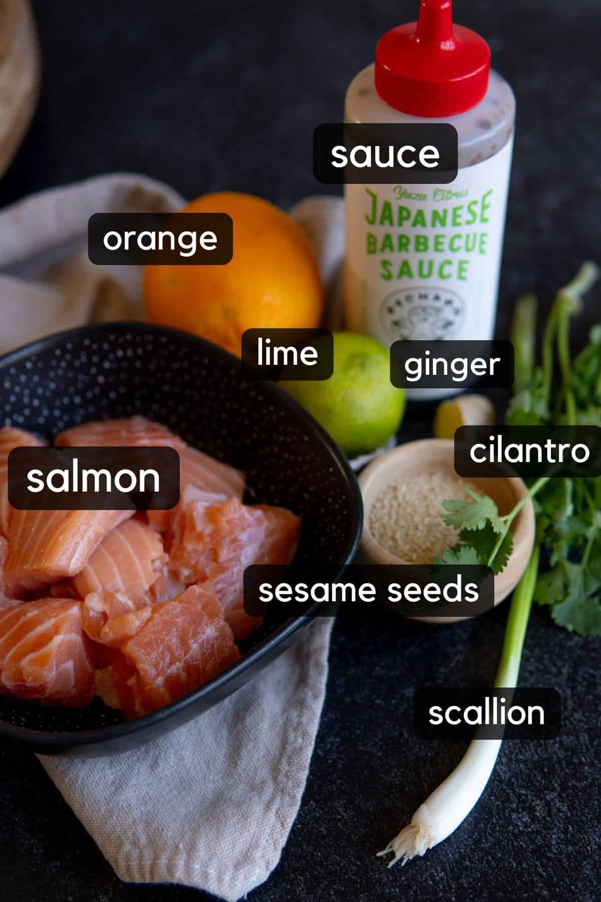 bowl with raw salmon chunks, orange next to it on table, lime, japanese barbecue sauce, ginger and other ingredients 