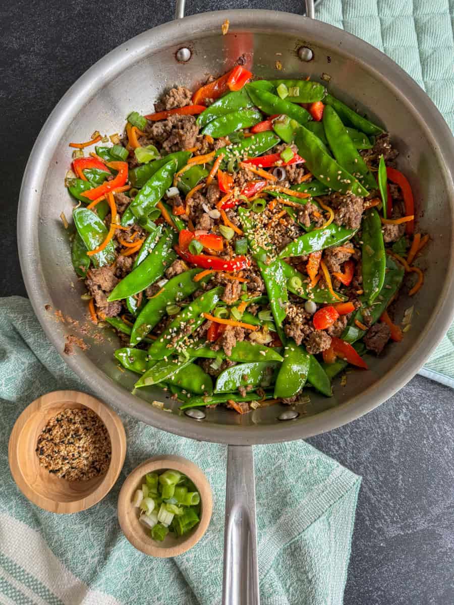 a large stainless steel pan with beef stir fry and snow peas and a side of sesame seeds and scallions 