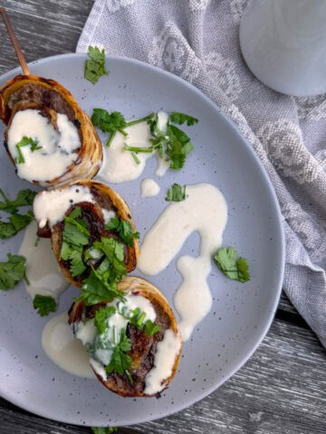 plate with a skewer and 3 beef roll ups with tahini sauce drizzled over it.
