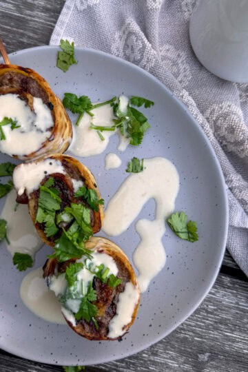 plate with a skewer and 3 beef roll ups with tahini sauce drizzled over it.