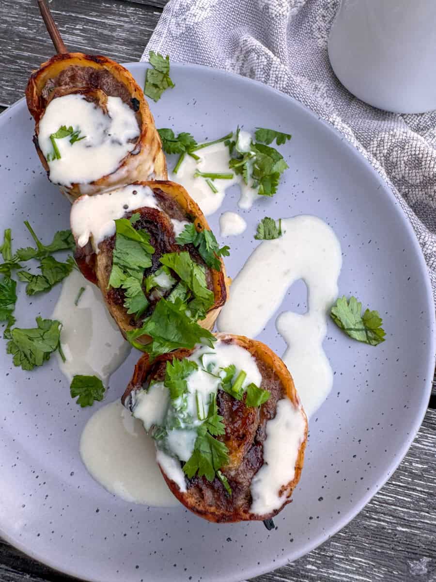closeup plate with 3 beef roll-ups on a skewer with tahini sauce drizzled on top. 