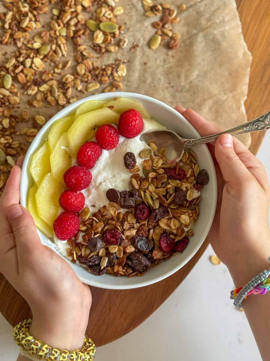 two child hands holding a bowl with yogurt, granola, kiwi and raspberries. 