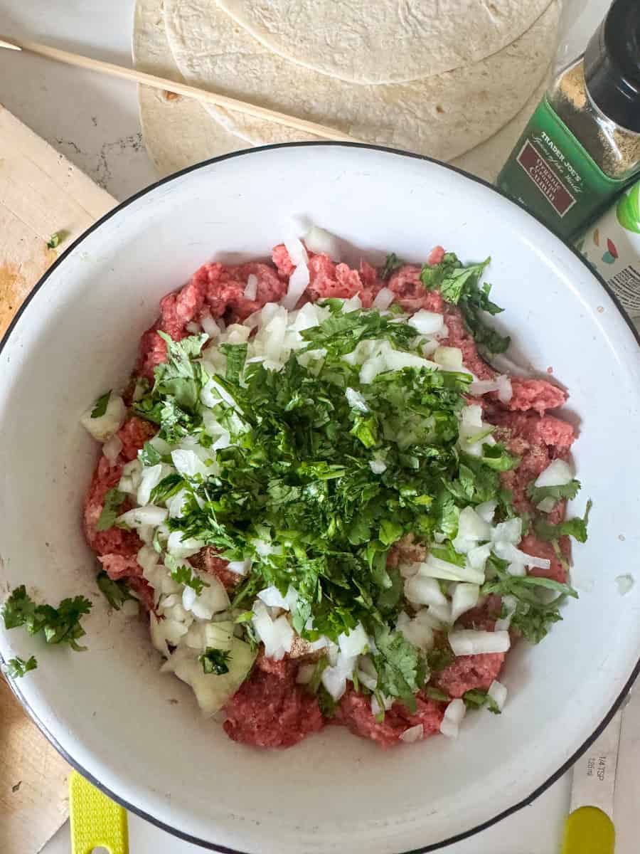 a bowl with ground beef, minced onion, and fresh green herbs. 