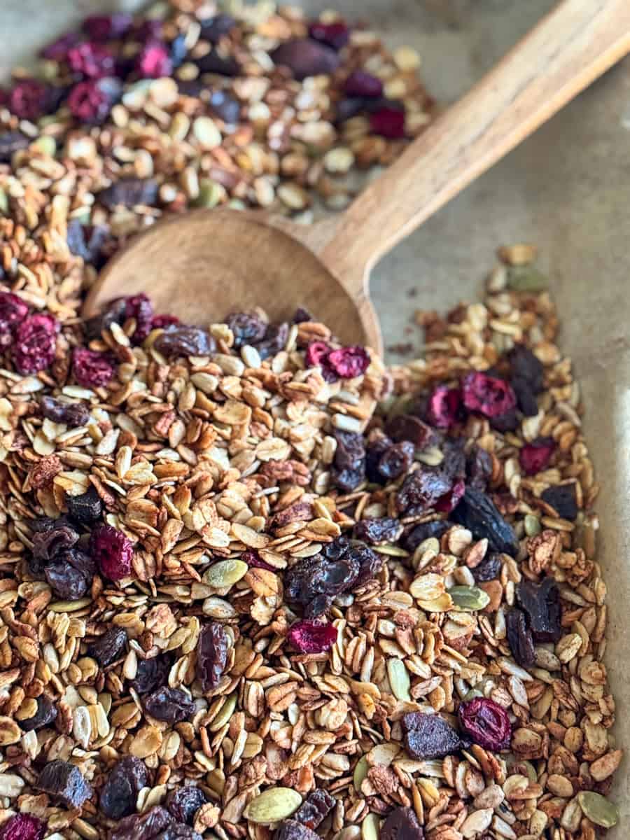 close up shot of parchment paper with toasted oats, raisins, cranberries, pumpkin seeds and a large wooden spoon.