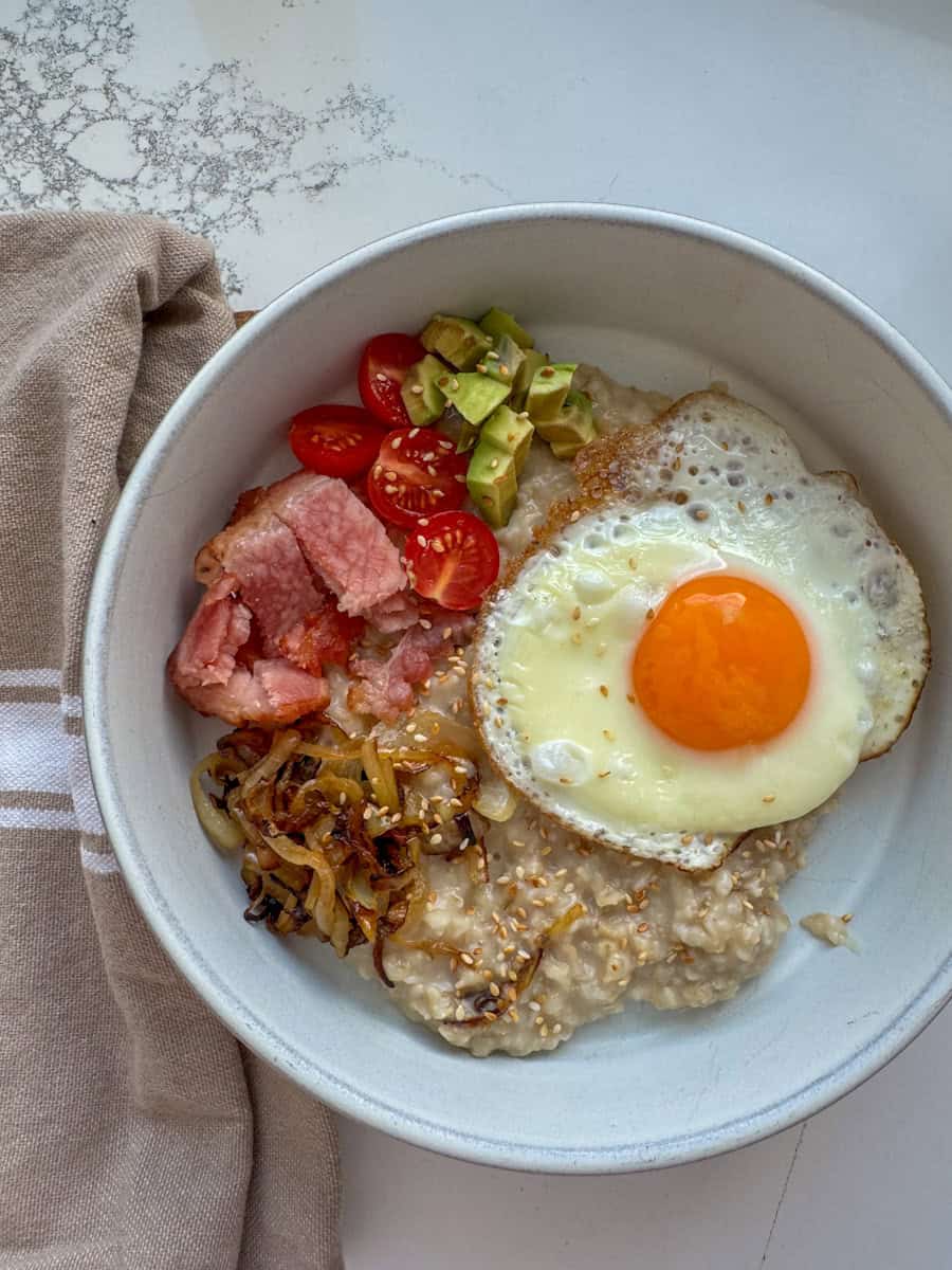bowl with steel cut oats, fried onion, canadian bacon, tomatoes, avocado, and sunny side up egg.